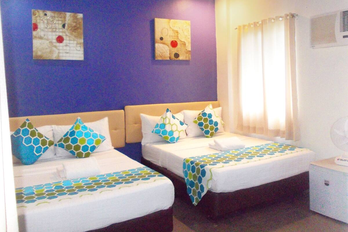A hotel room with two beds, each with a patterned bedspread and matching pillows. There is a ceiling fan, a window with curtains, and artwork on the walls.