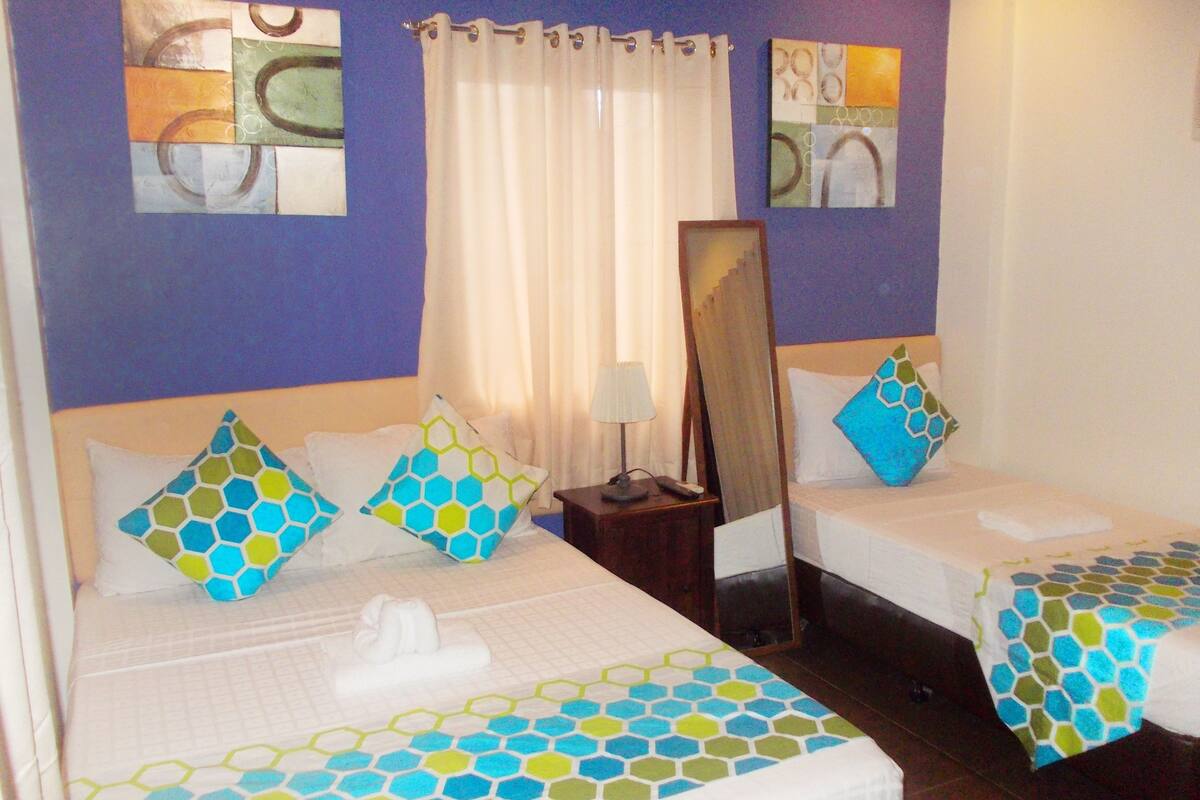 A hotel room with two beds, each with a white bedspread and matching pillows. There is a nightstand with a lamp and a mirror between the beds. The room has a purple accent wall and artwork above the beds.
