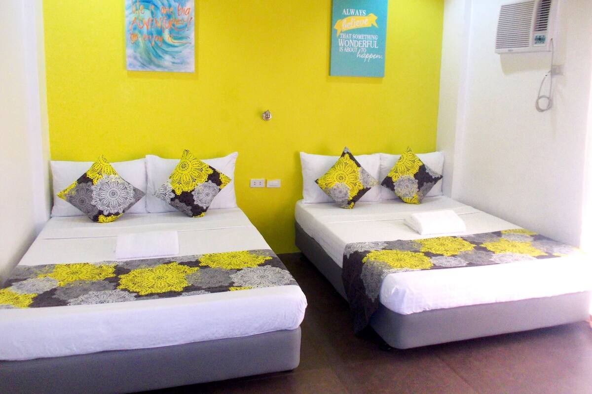 A room with two beds, each with a white mattress and a blanket with a yellow and gray pattern. There are two pillows on each bed, one with a yellow and gray pattern and the other with a gray and white pattern. The walls are yellow, and there are two framed quotes on the wall.