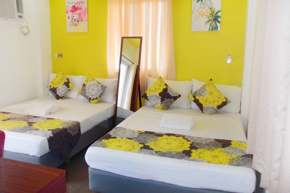 A room with two beds, each with a white mattress and a patterned blanket. There are decorative pillows on the beds, and a mirror on the wall. The room has a yellow wall and a window with curtains.