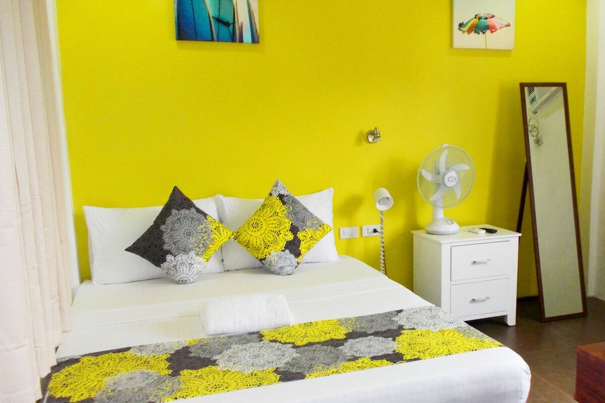 A bed with a white and gray blanket, two pillows with a yellow and gray pattern, a white nightstand with a lamp, a mirror, and a fan.