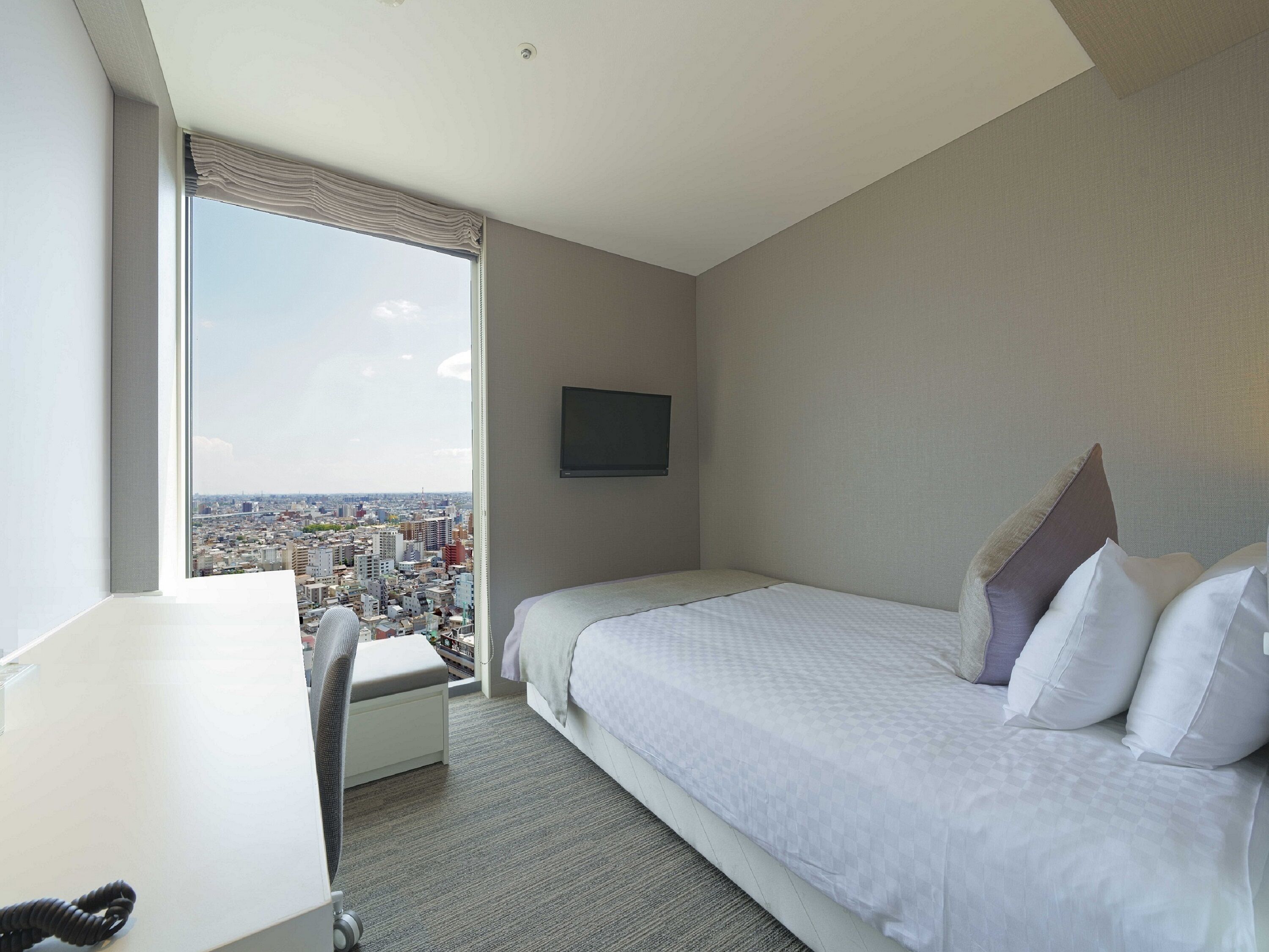 A hotel room with a bed, a desk, a chair, a TV, and a large window offering a city view.