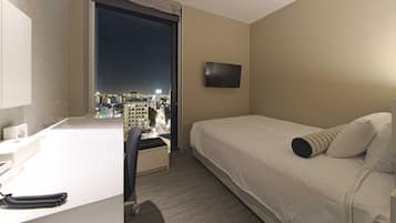 Una camera d'albergo con un letto, una scrivania, una sedia, una televisione e un'ampia finestra con vista sulla città.