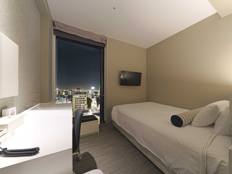 A hotel room with a bed, a desk, a chair, a TV, and a large window offering a city view.