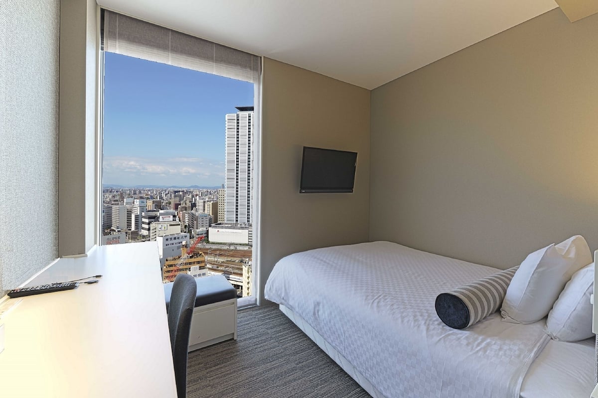 A hotel room with a bed, a desk, and a large window offering a city view.
