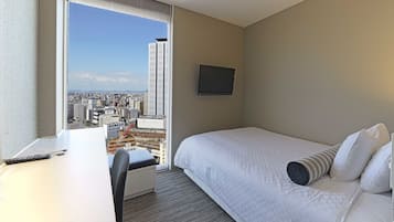 Quarto de hotel com cama, escrivaninha e uma grande janela com vista para a cidade.