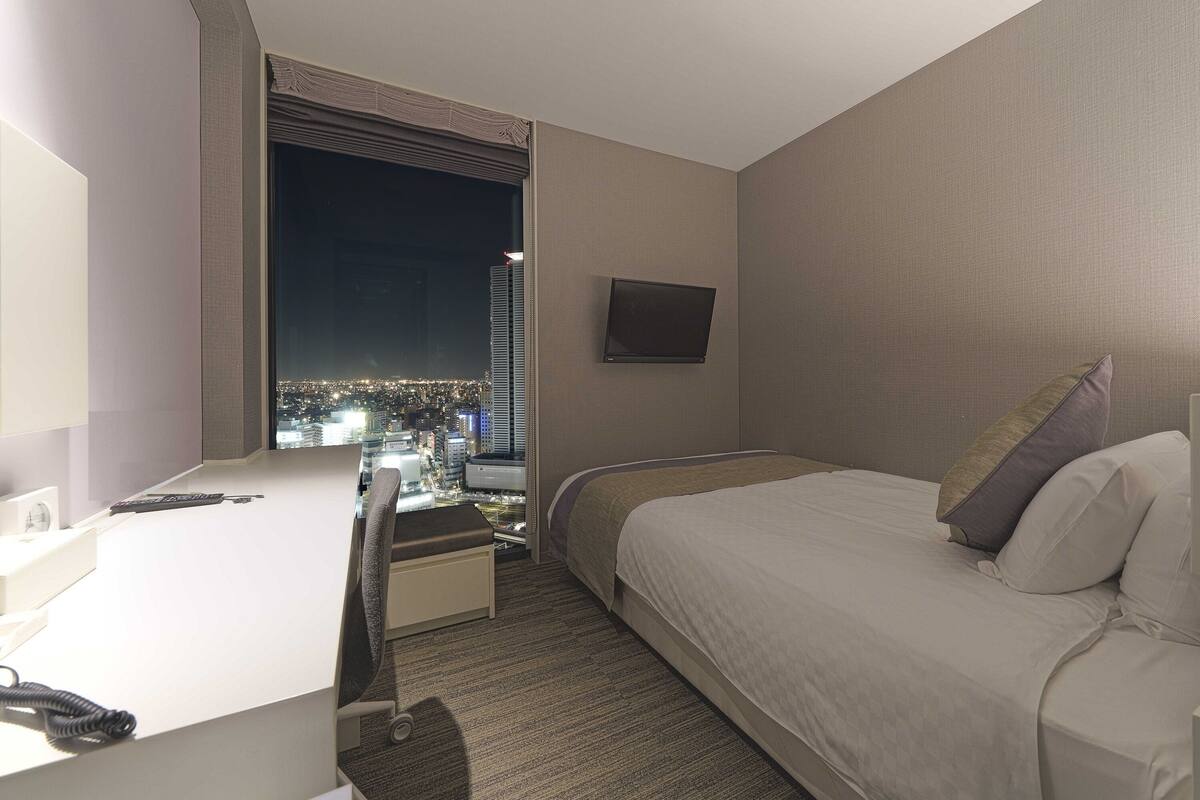 A hotel room with a bed, a desk, a chair, a TV on the wall, and a view of a cityscape at night.