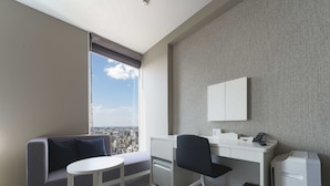 Una camera d'albergo moderna con un divano grigio, un piccolo tavolino rotondo, una scrivania con una sedia, un armadio bianco e vista sulla città da una grande finestra.