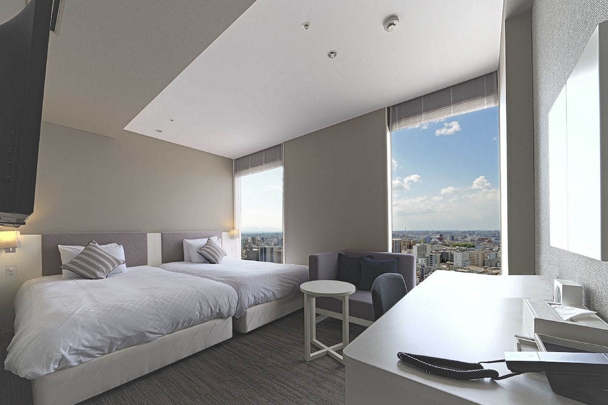 A modern hotel room with two beds, a small seating area, a desk, and a large window offering a city view.