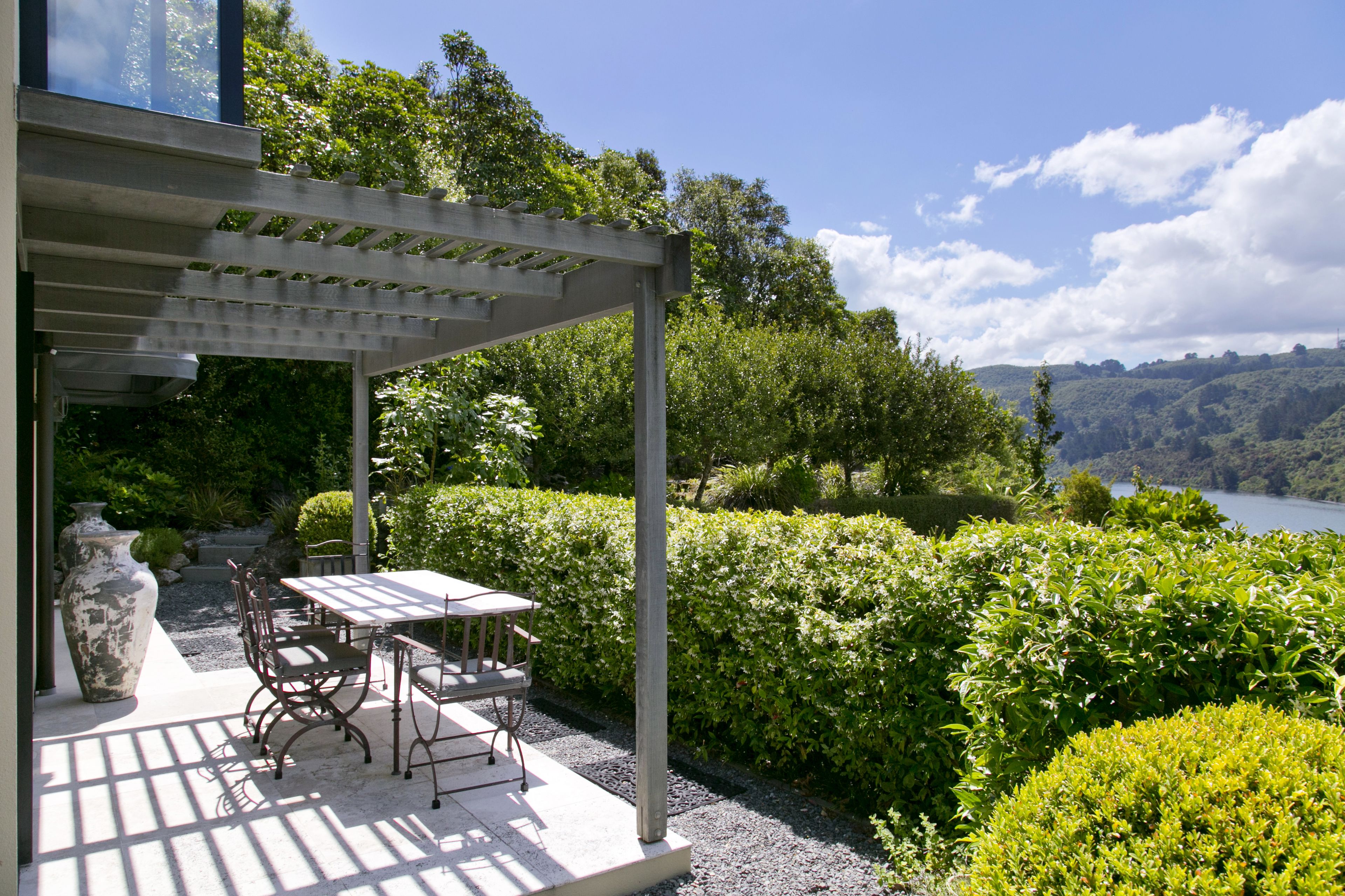 A covered patio with a table and chairs, surrounded by lush greenery and a view of a lake.