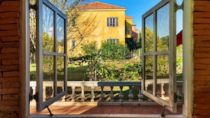 An open arched window with a stone wall base, revealing a yellow building with a red roof and a garden in the background. - Davinci Di Khao Yai (Pak Chong)