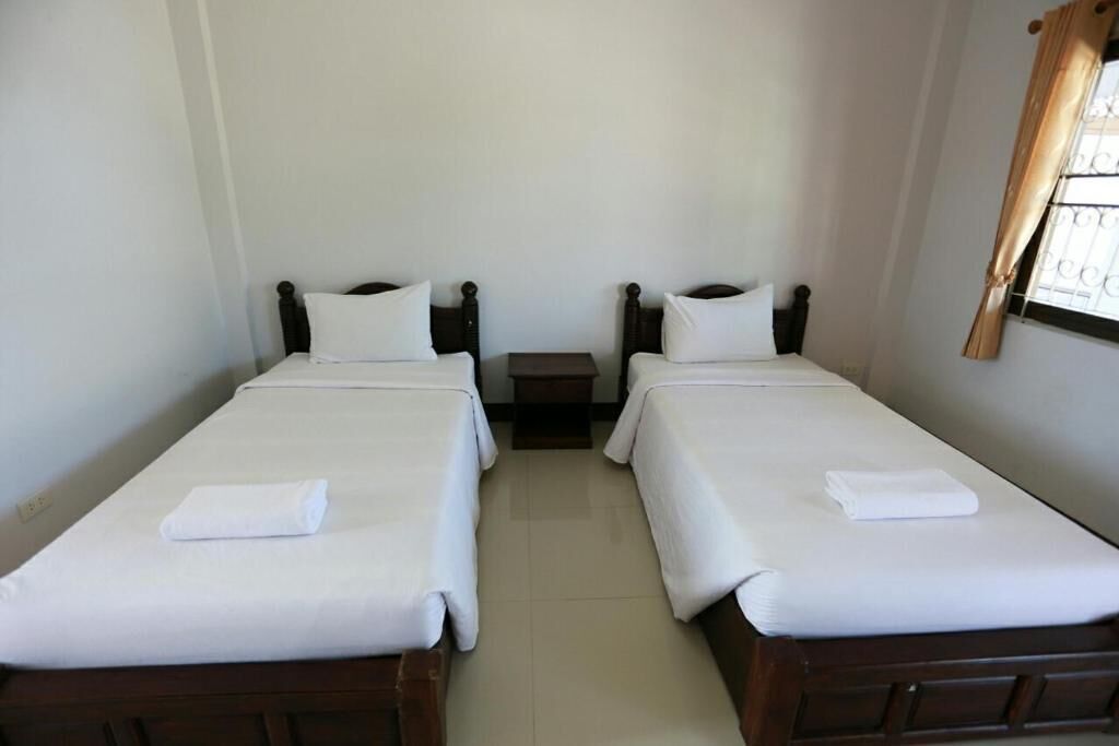 A room with two single beds, each with a wooden headboard and white bedding. There is a small bedside table between the beds, and a window with curtains on the right side.