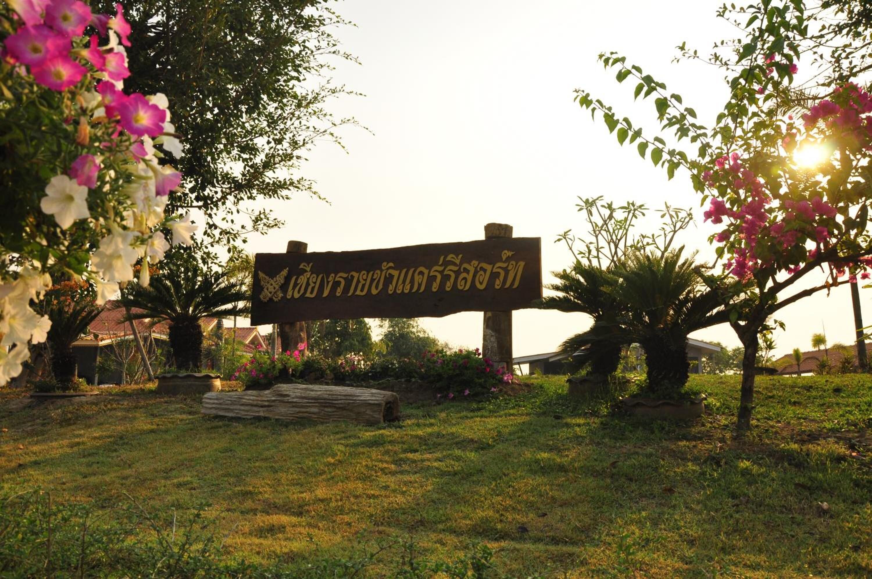 A sign with Thai script in a garden setting with flowering plants and a wooden bench.