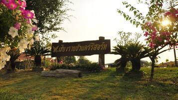 A sign with Thai script in a garden setting with flowering plants and a wooden bench.