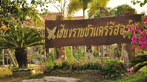 A wooden sign with Thai script, surrounded by lush greenery and flowering plants.