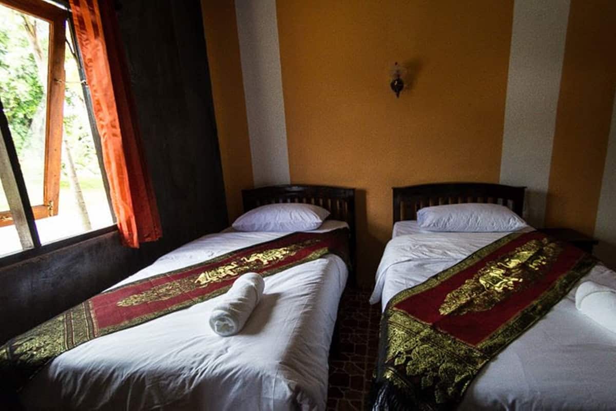 Two single beds with white linens and red and gold decorative bedspreads, a window with curtains, and a wall-mounted light fixture.