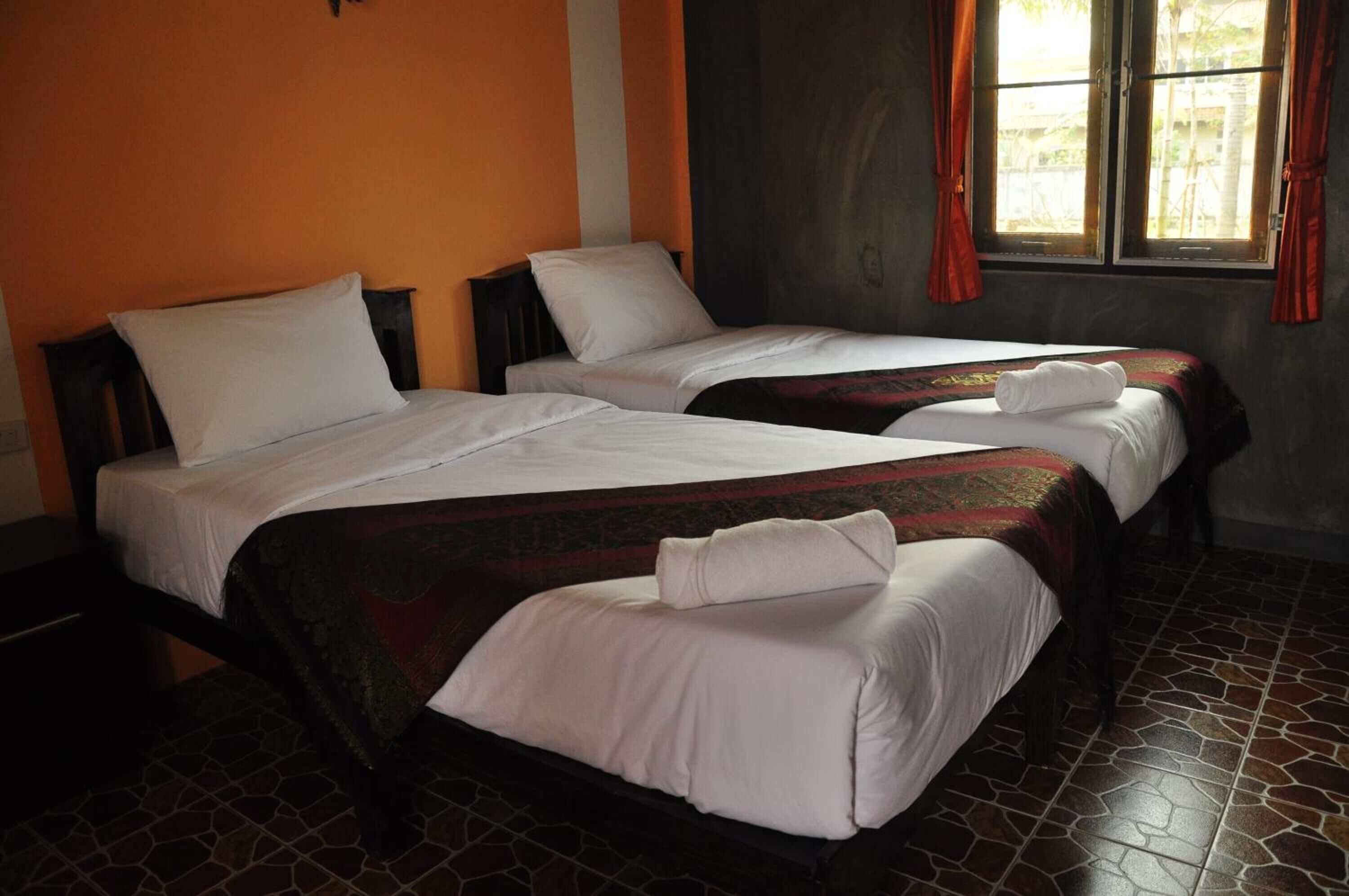 Dos camas individuales con sábanas blancas y cabeceros oscuros en una habitación con paredes anaranjadas y una ventana con cortinas rojas.