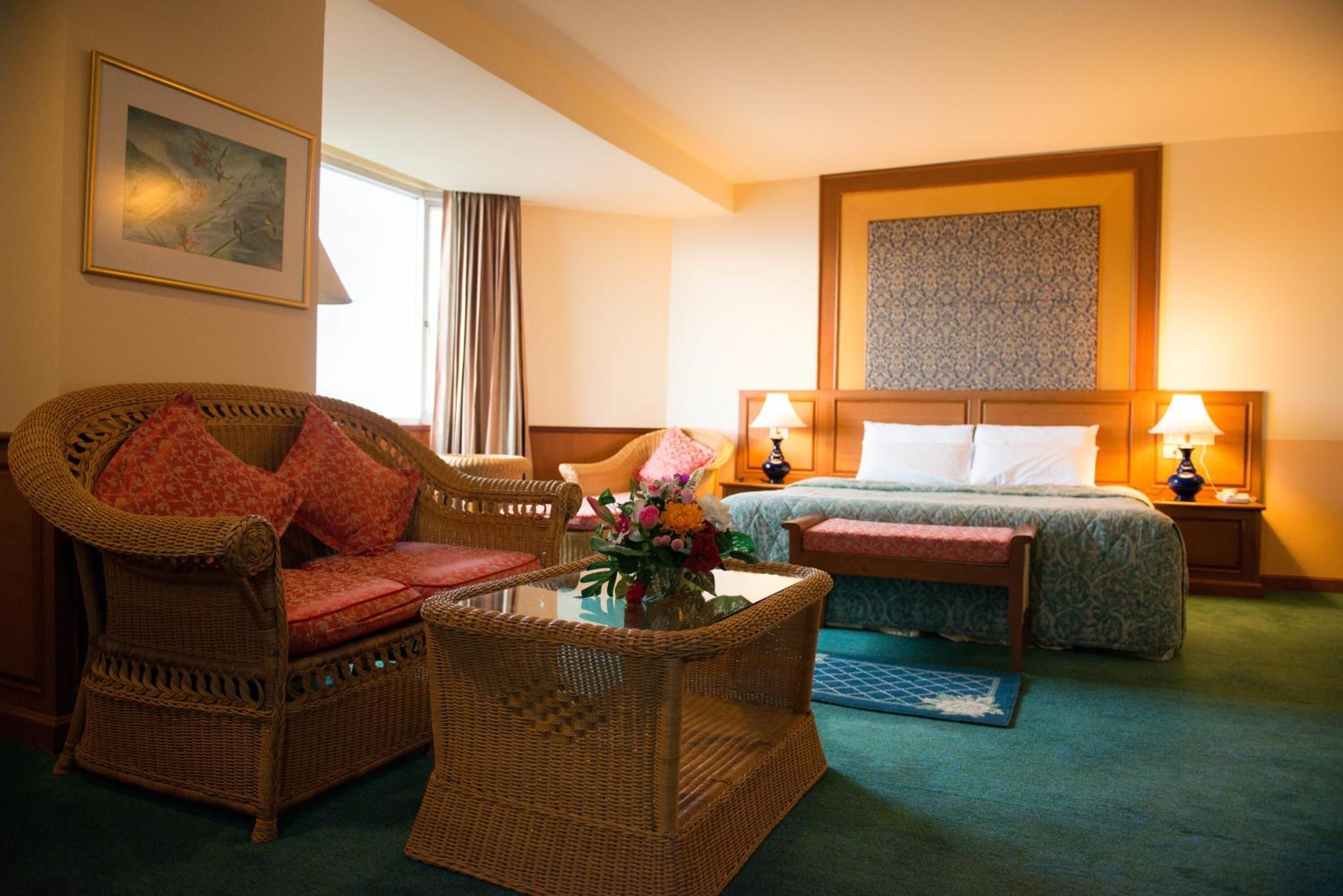 A hotel room with a wicker sofa, a bed with a patterned headboard, a bedside table with lamps, and a framed picture on the wall.
