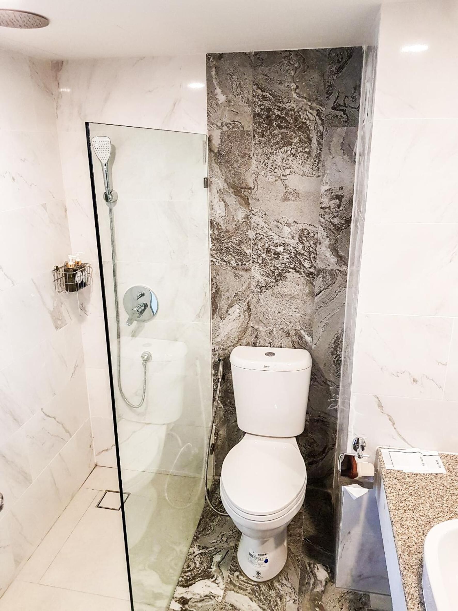Un baño con ducha envuelta en vidrio, inodoro blanco y pared con diseño de mármol.