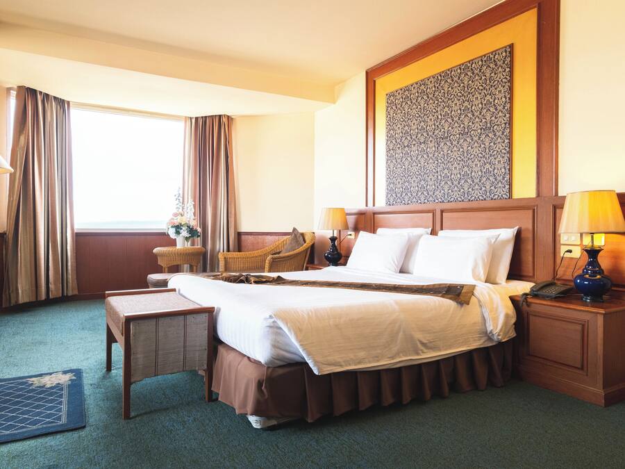 A hotel room with a large bed, two bedside tables, a nightstand, a lamp, a chair, and a view of the outdoors.