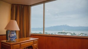 Una camera d'albergo con una scrivania in legno, una lampada e un'ampia finestra che offre vista su un corpo d'acqua e montagne in lontananza.
