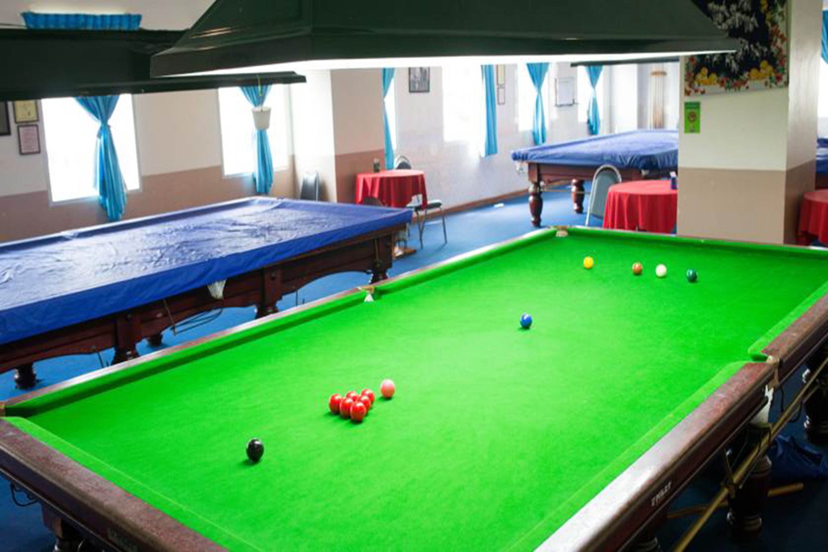 Uma mesa de bilhar com bolas, mesa vermelha e mesa azul em um ambiente com janelas e cortinas.