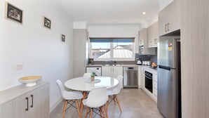 A modern kitchen with white cabinets, a stainless steel refrigerator, a dining table with white chairs, and a window providing natural light.