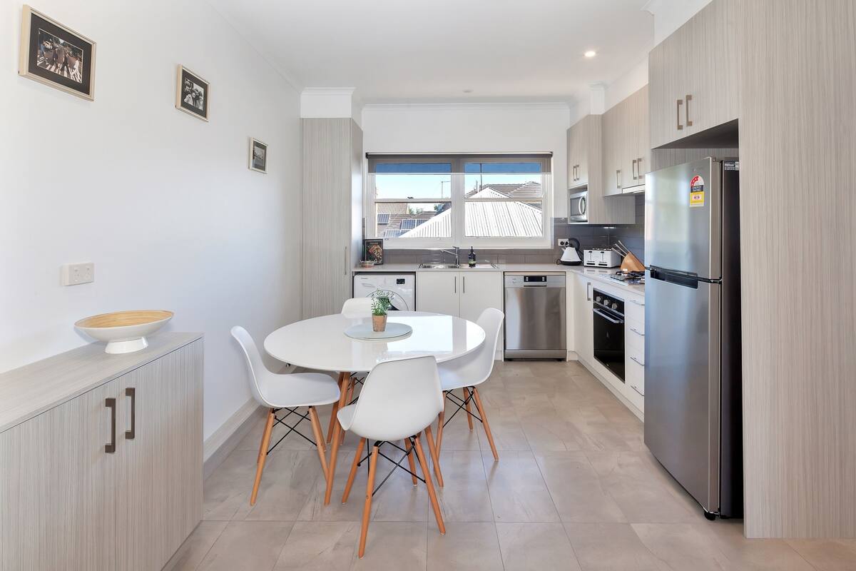 Una cocina moderna con armarios blancos, un refrigerador de acero inoxidable, una mesa de comedor con sillas blancas y un ventanal que proporciona luz natural.