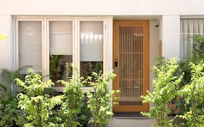A building with a wooden door, a window with a metal grill, and a small porch area with potted plants. - Benri sriracha residence (Si Racha)