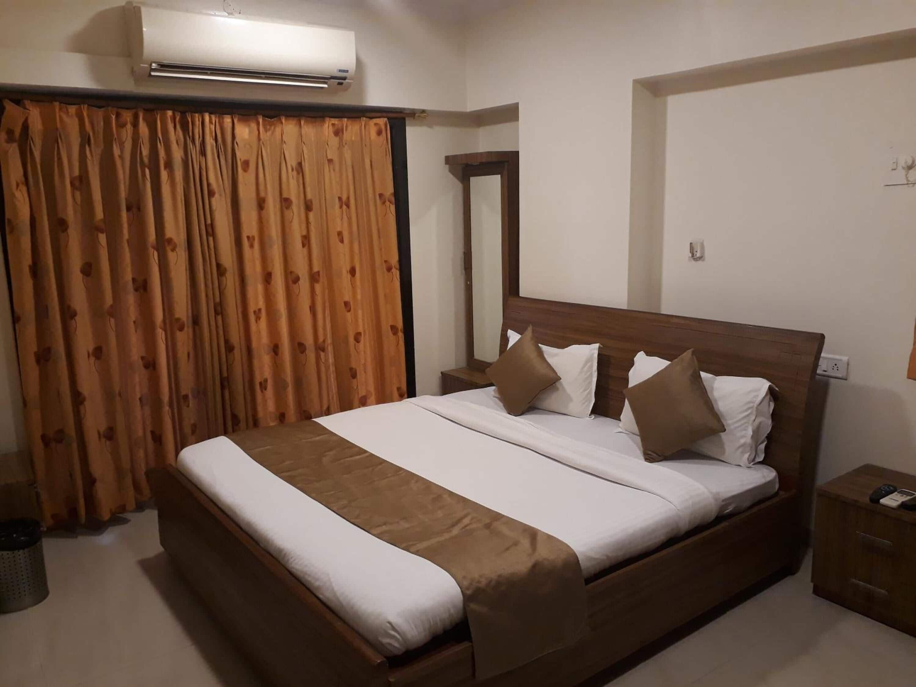 A hotel room with a large bed, brown curtains, an air conditioning unit, a wooden nightstand, and a mirror.