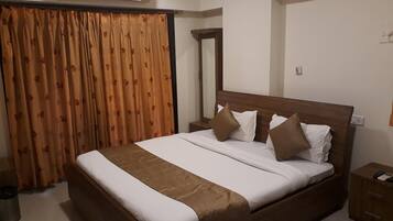A hotel room with a large bed, brown curtains, an air conditioning unit, a wooden nightstand, and a mirror.