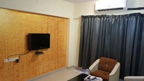 Habitación con televisor de pantalla plana montado en una pared texturizada, un sofá, una mesa de centro con revistas y un ventanal con cortinas oscuras.