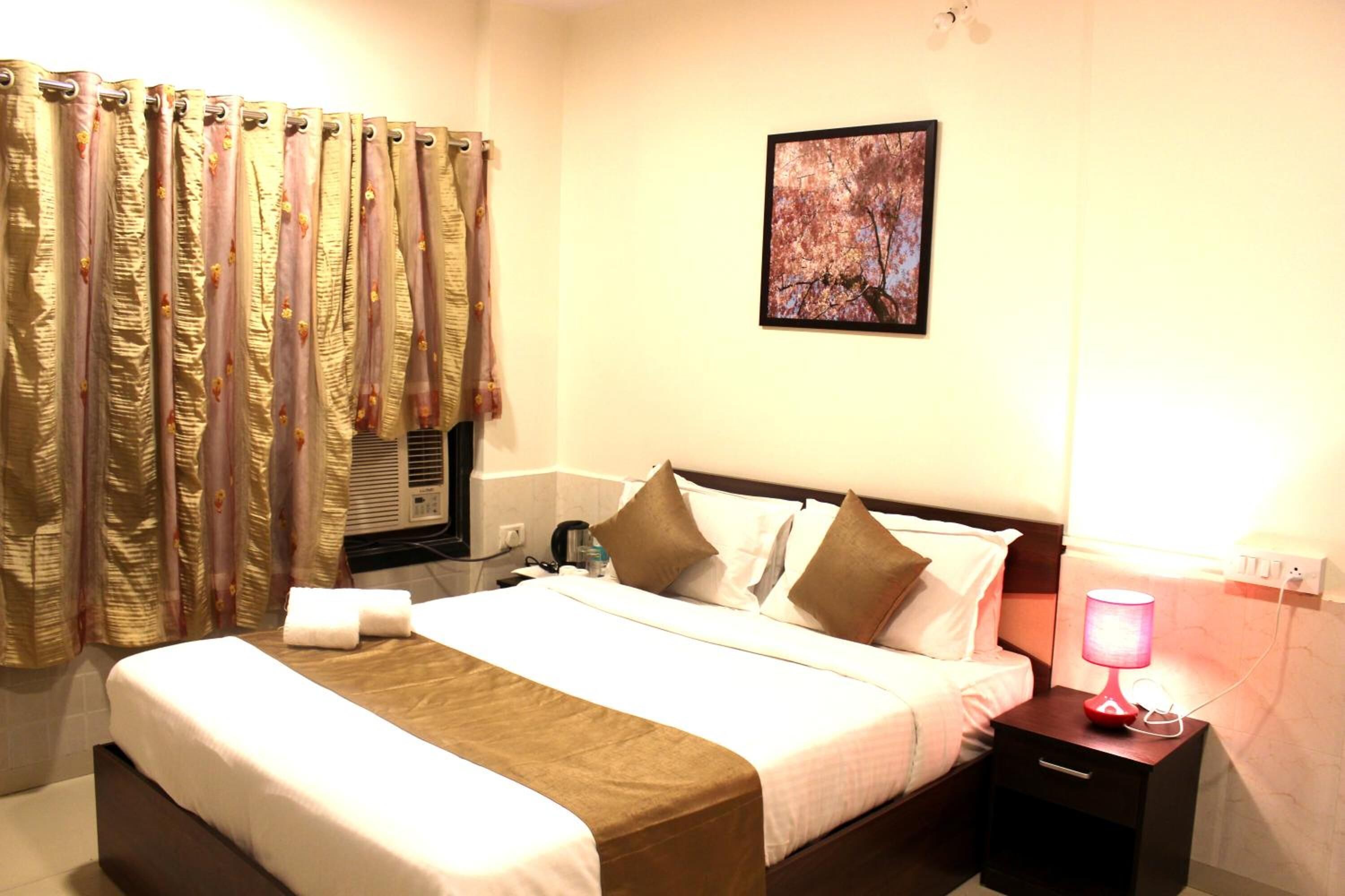 Habitación de hotel con cama, mesita de noche con lámpara, ventana con cortinas e imagen colgada en la pared.