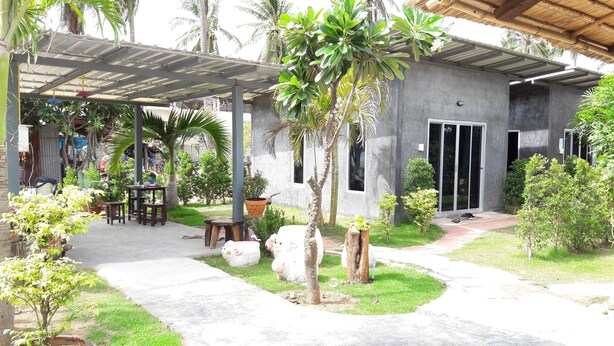 A covered outdoor area with a concrete floor, surrounded by greenery and palm trees. There are outdoor seating arrangements and a small statue.