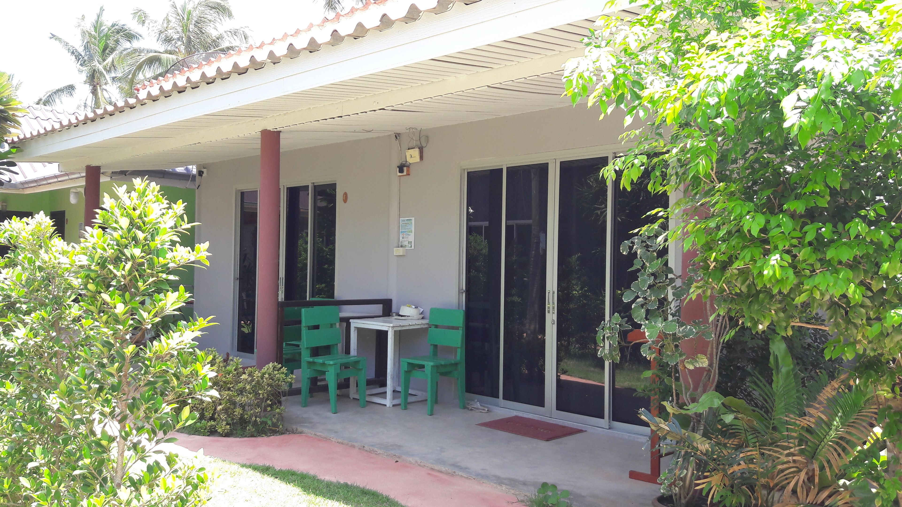 A covered outdoor seating area with a table and chairs, a door, and a small garden with plants.