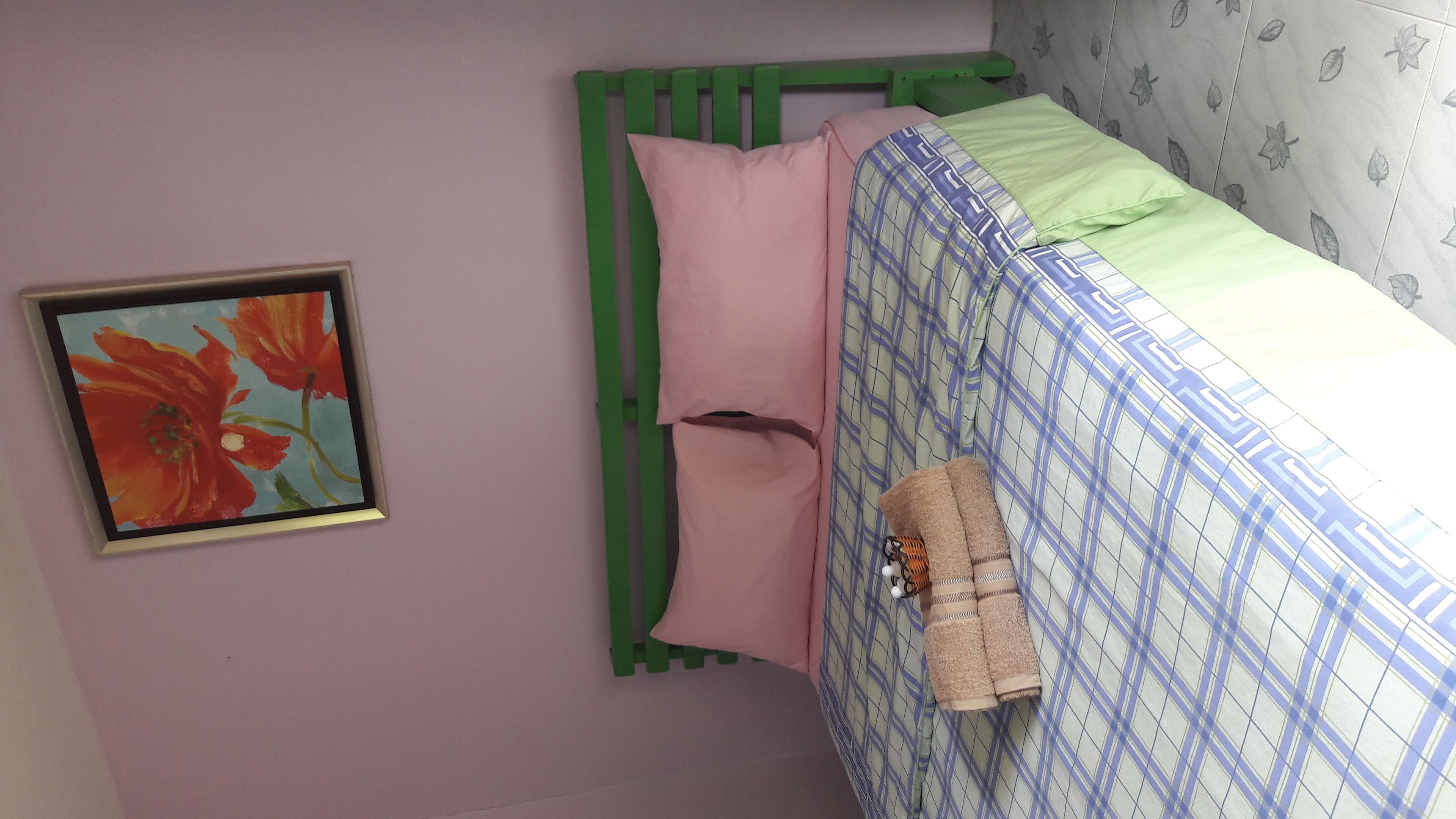 Un dormitorio con un marco de cama metálico verde, una almohada rosa y una colcha blanca y azul a cuadros.