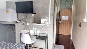 A compact hotel room with a bed, a small desk with a chair, a flat-screen TV mounted on the wall, a refrigerator, and a door with a wooden panel. - Hive28 (Bangkok)