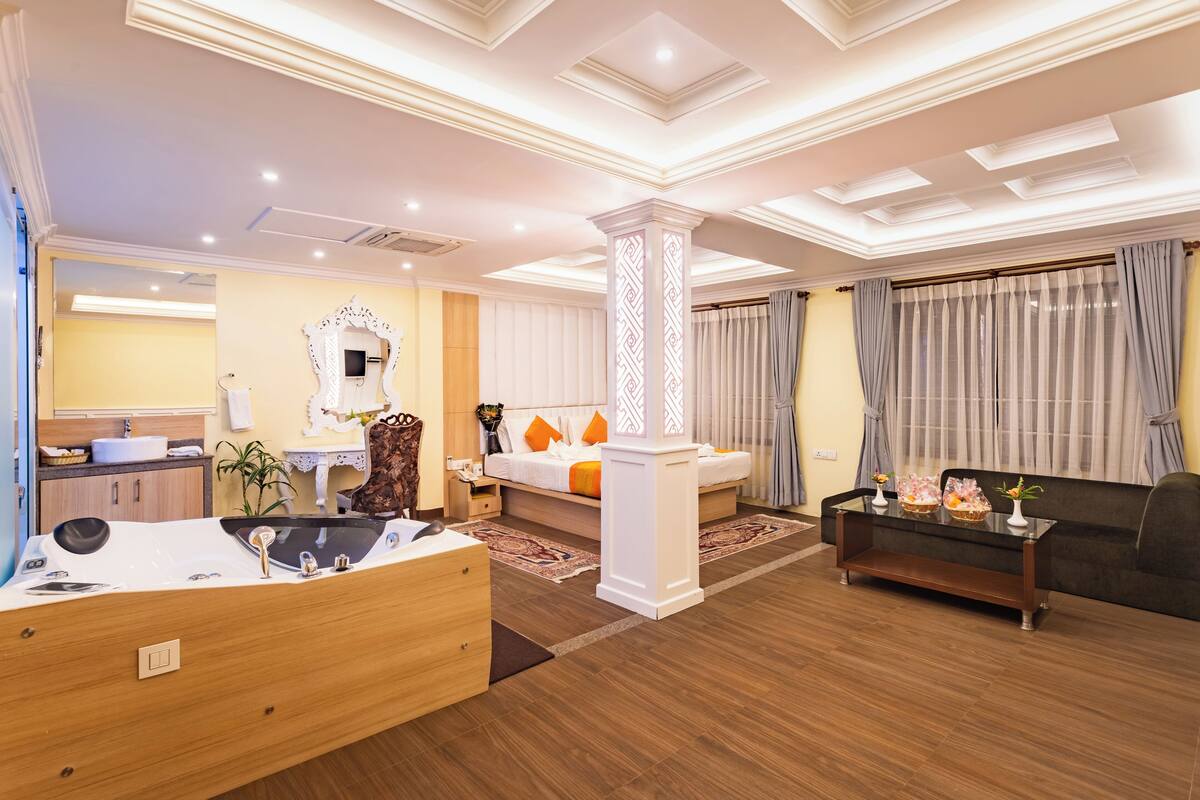 Una habitación de hotel moderna con suelo de madera, un área de estar con sofá y mesa de centro, un espejo grande y una mesa de comedor con sillas.