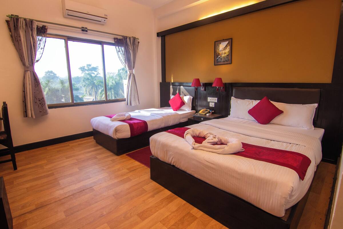 A hotel room with two beds, a wooden floor, a window with curtains, a nightstand with a lamp, and a painting on the wall.