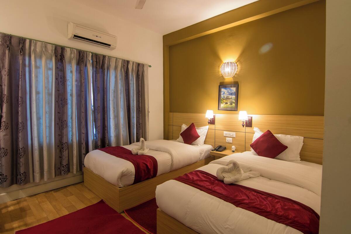 A hotel room with two beds, a red carpet, an air conditioning unit, a wall-mounted light fixture, and a painting above the bed.