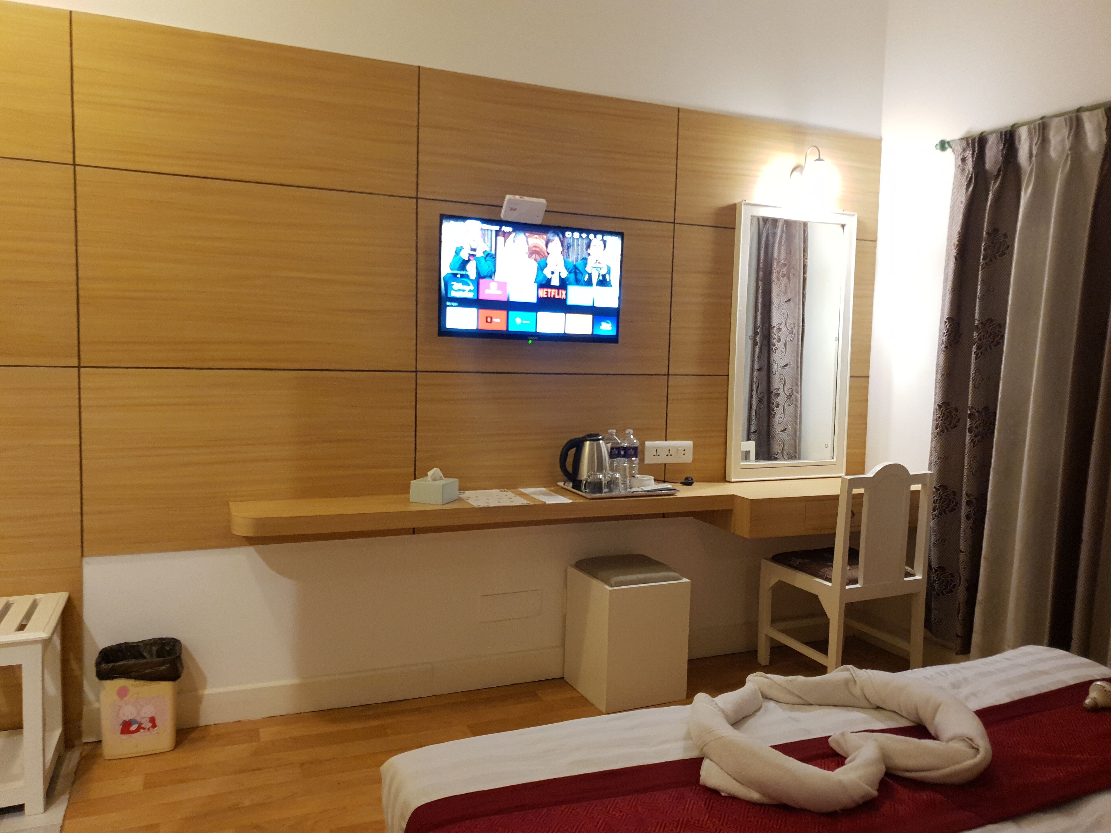 A hotel room with a wooden panel wall, a flat-screen TV mounted above a wooden desk, a small trash bin, a bedside table, and a bed with a red blanket.