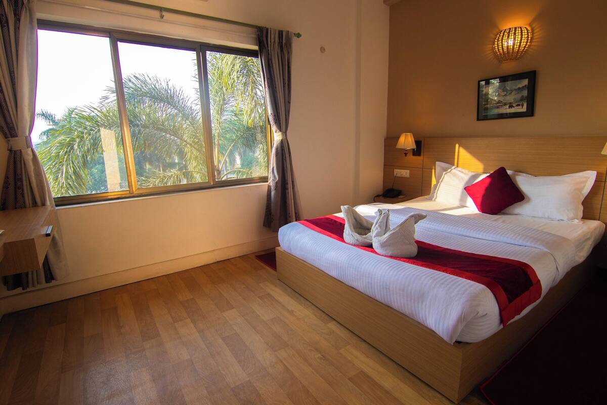 A hotel room with a wooden floor, a large bed with white bedding and red accents, a nightstand with a lamp, and a window with curtains.