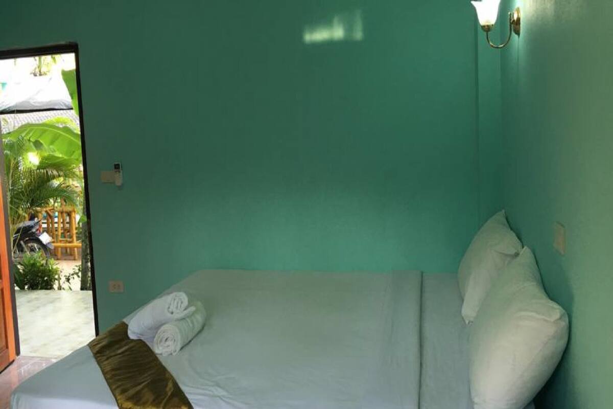 A single bed with white linens and a gold accent, a teal wall, and a window with a view of a patio area.