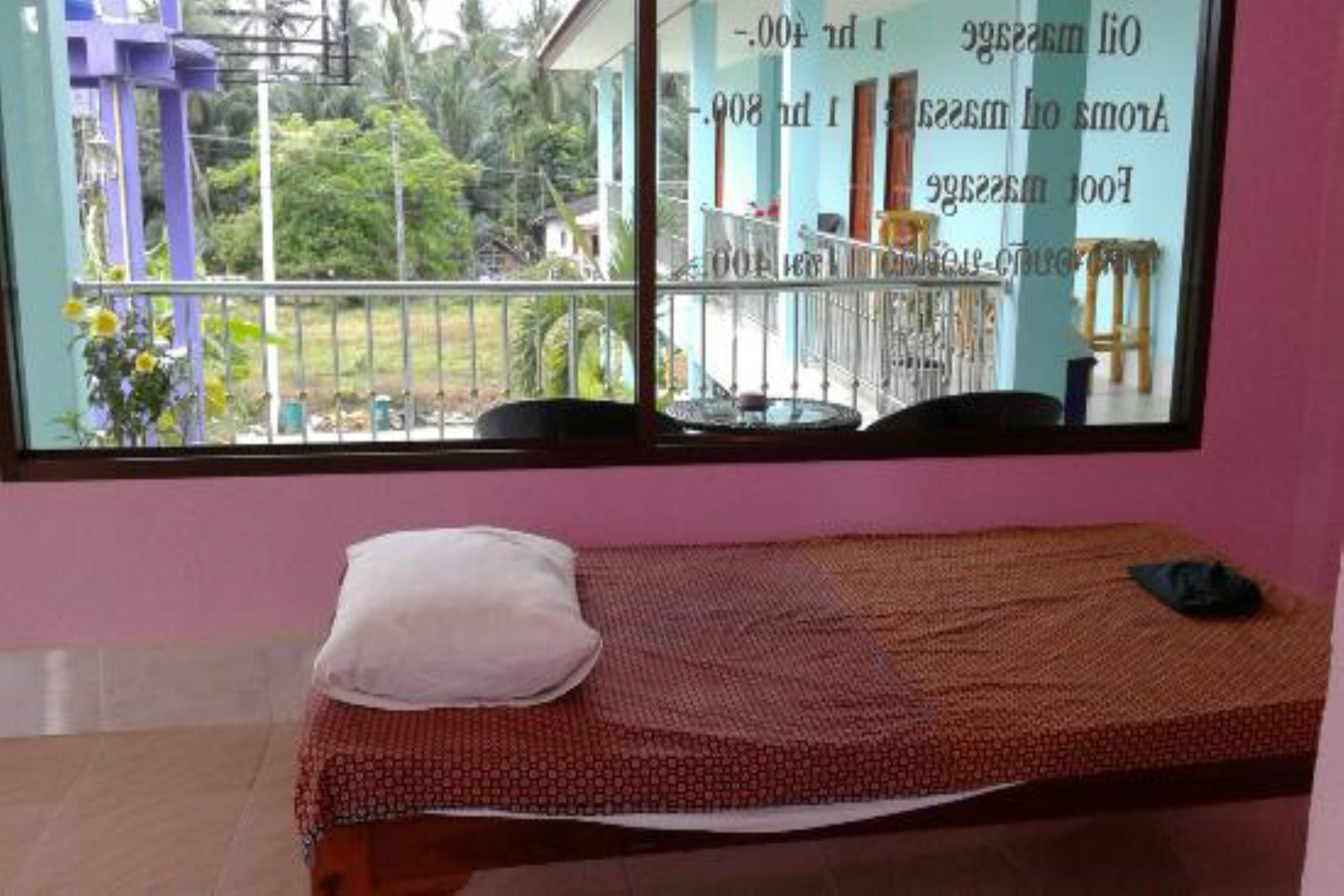 A single bed with a patterned bedspread and a pillow, a window with a view of a balcony and greenery, and a pink wall.