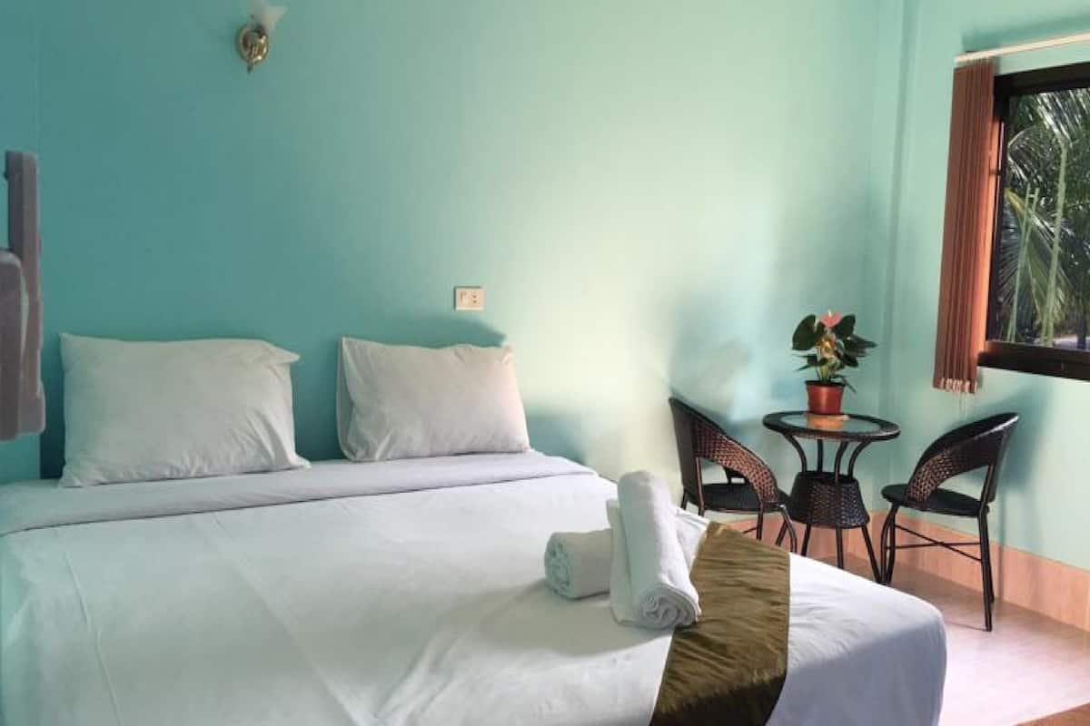 A hotel room with a bed, two pillows, a rolled towel, a small table with chairs, and a window with a view of palm trees.