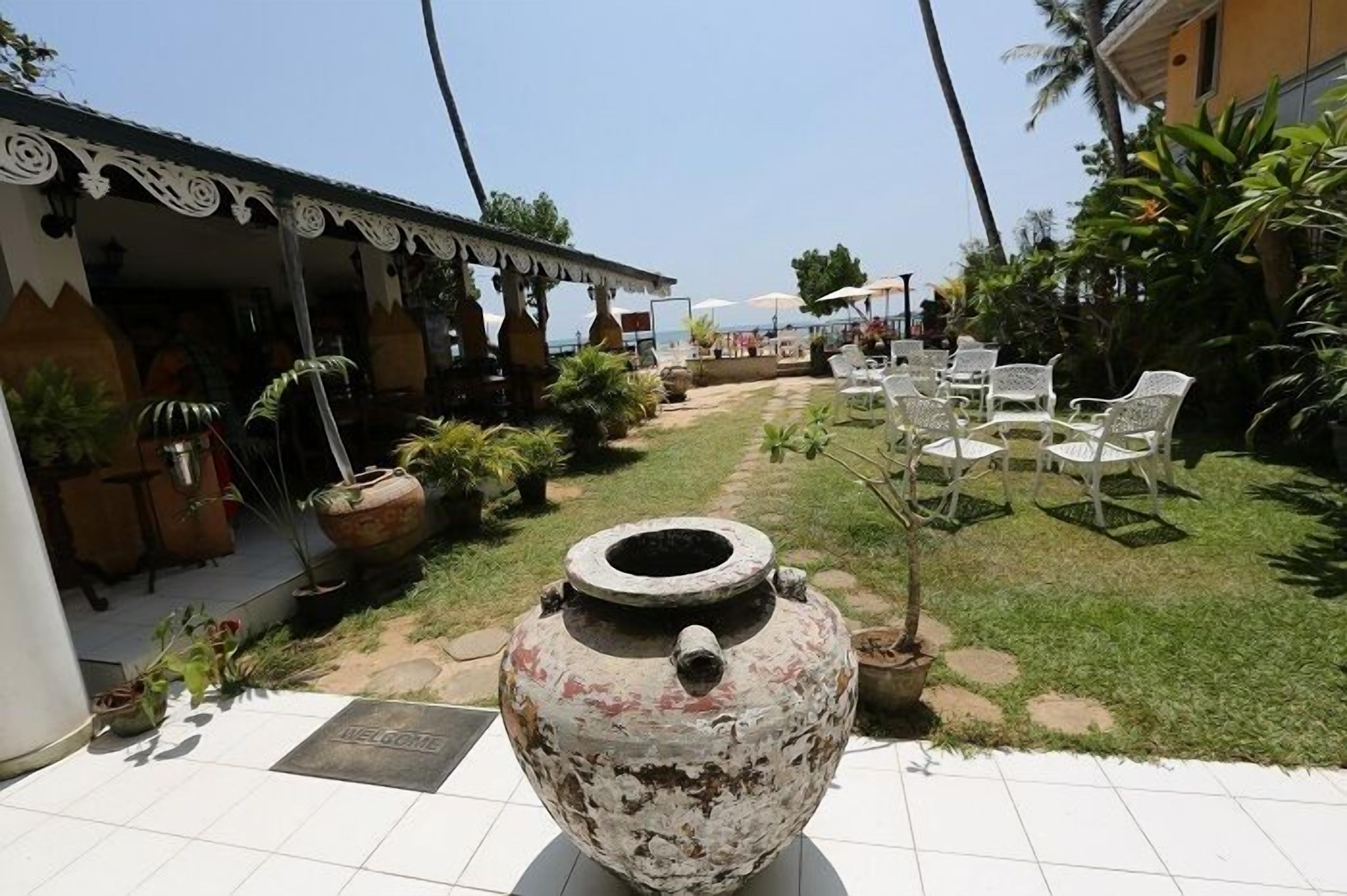 Un espace extérieur aménagé avec des chaises et des tables blanches, entouré de plantes en pot et agrémenté d’un grand pot décoratif.