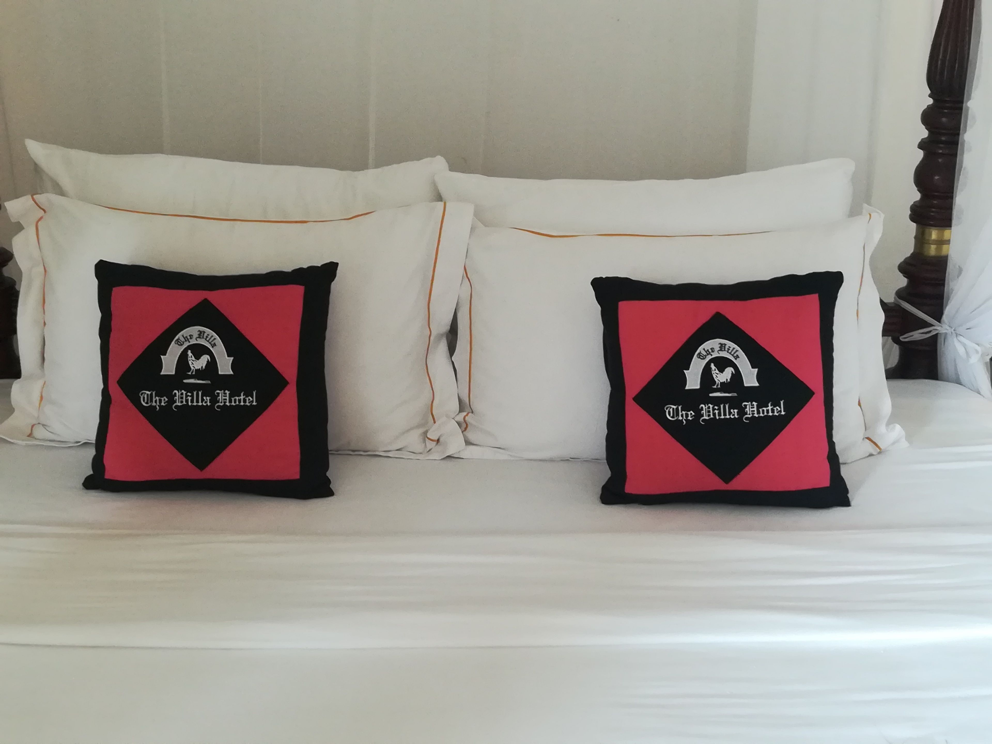 Un lit avec une literie blanche et deux coussins décoratifs à motifs noirs et rouges, représentant un coq et l’inscription « The Villa Hotel ».