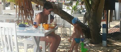 Un área de comedor frente al mar con sillas y mesas de plástico blanco, un tronco de árbol grande y un niño jugando en la arena.