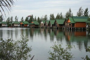 A row of wooden cabins with green roofs, situated on stilts over a calm body of water, surrounded by trees. - Khungkapong Resort (Phetchaburi)