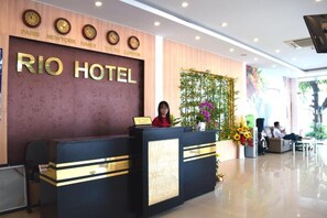 Un mostrador de recepción de hotel con una recepcionista, un reloj que muestra la hora en París, Nueva York, Hanoi, Tokio y Londres, y un cartel grande que dice "RIO HOTEL".